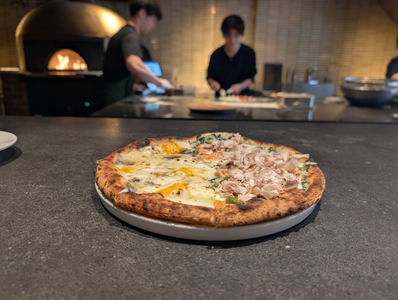 石窯の炎が奥で輝くオープンキッチンを背景に、グレーのカウンター上に置かれた焼きたてのハーフ＆ハーフピザ